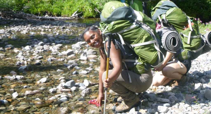 two teens wearing backpacks kneel beside a rocky stream on a outward bound course